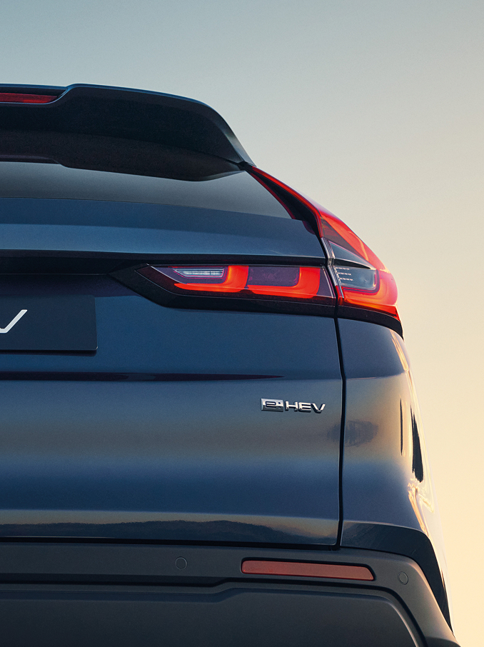 Detailný pohľad zozadu na tmavomodrú Honda CR-V e:HEV s logom Honda, nápisom CR-V a osvetlenými charakteristickými zadnými LED svetlami na pozadí oblohy pri západe slnka.
