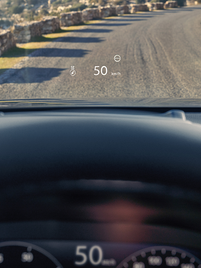 Pohľad z miesta vodiča, kde Head-Up Display (HUD) zobrazuje „50 km/h“ a kompas na čelné sklo nad štrkovou cestou.