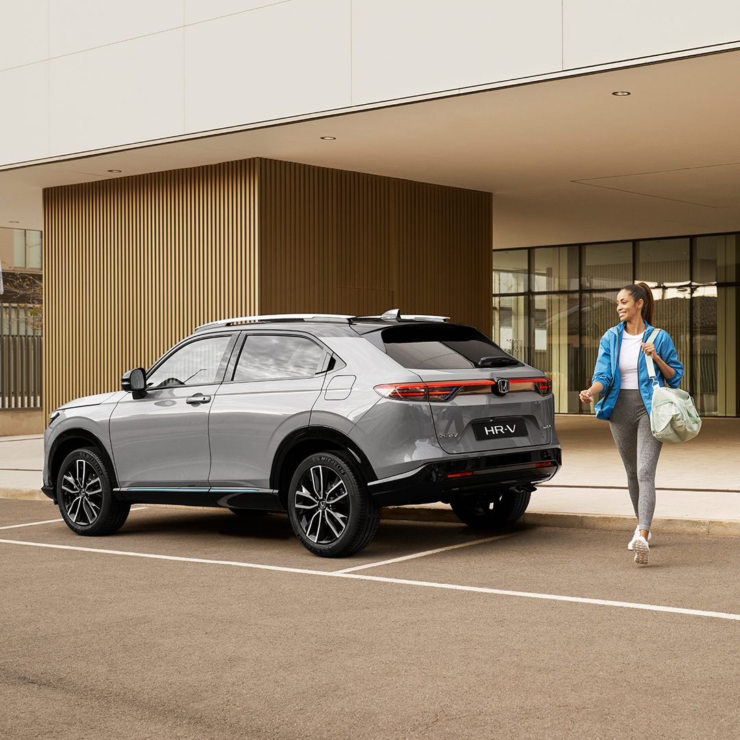 Strieborná Honda HR-V zaparkovaná pred modernou budovou, zatiaľ čo okolo nej prechádza žena s taškou, čo naznačuje každodenné mestské využitie a komfort.