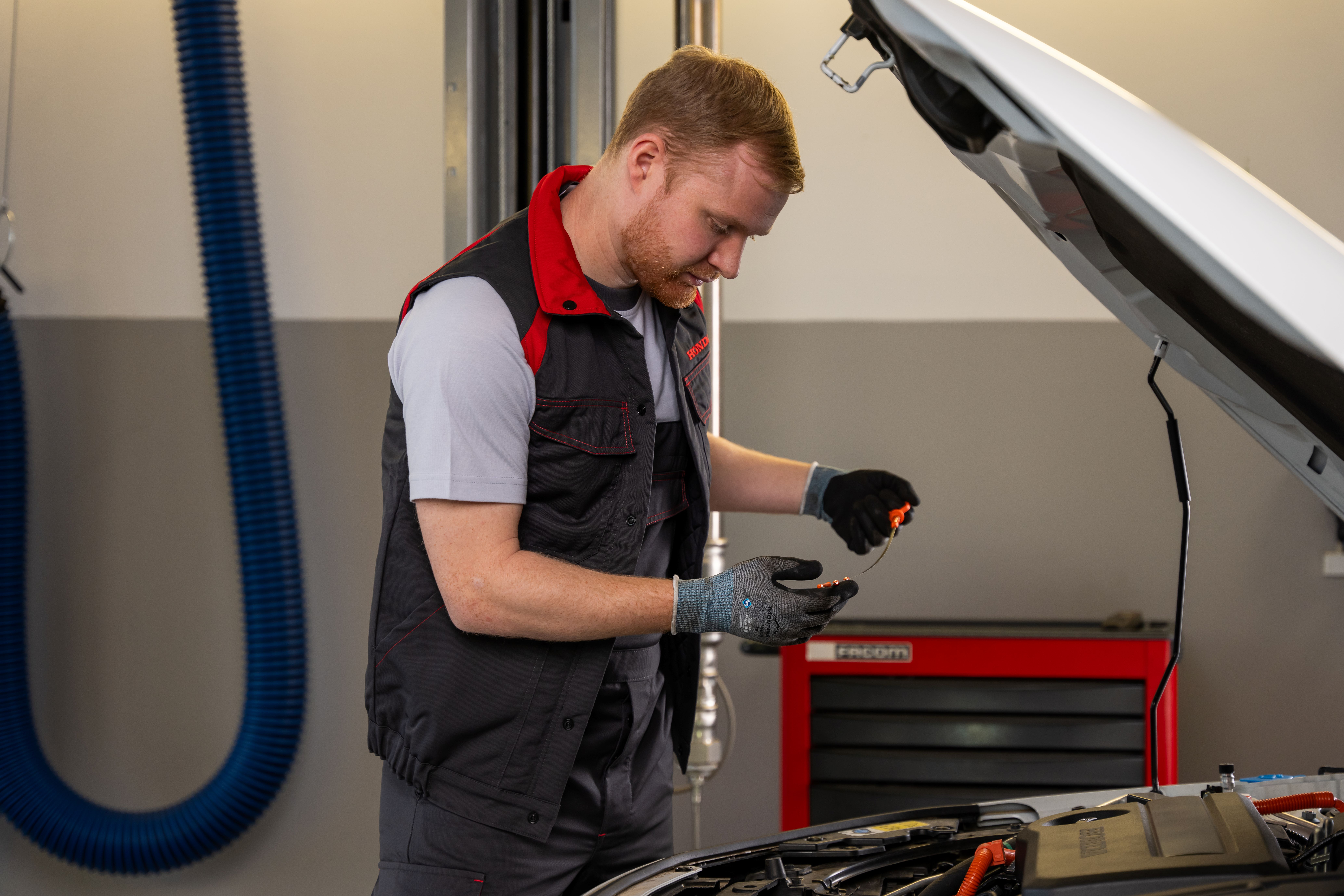 A mechanic in a red and gray uniform works on a Honda vehicle with an open hood. A red toolbox is visible beside him in what appears to be an auto repair shop.