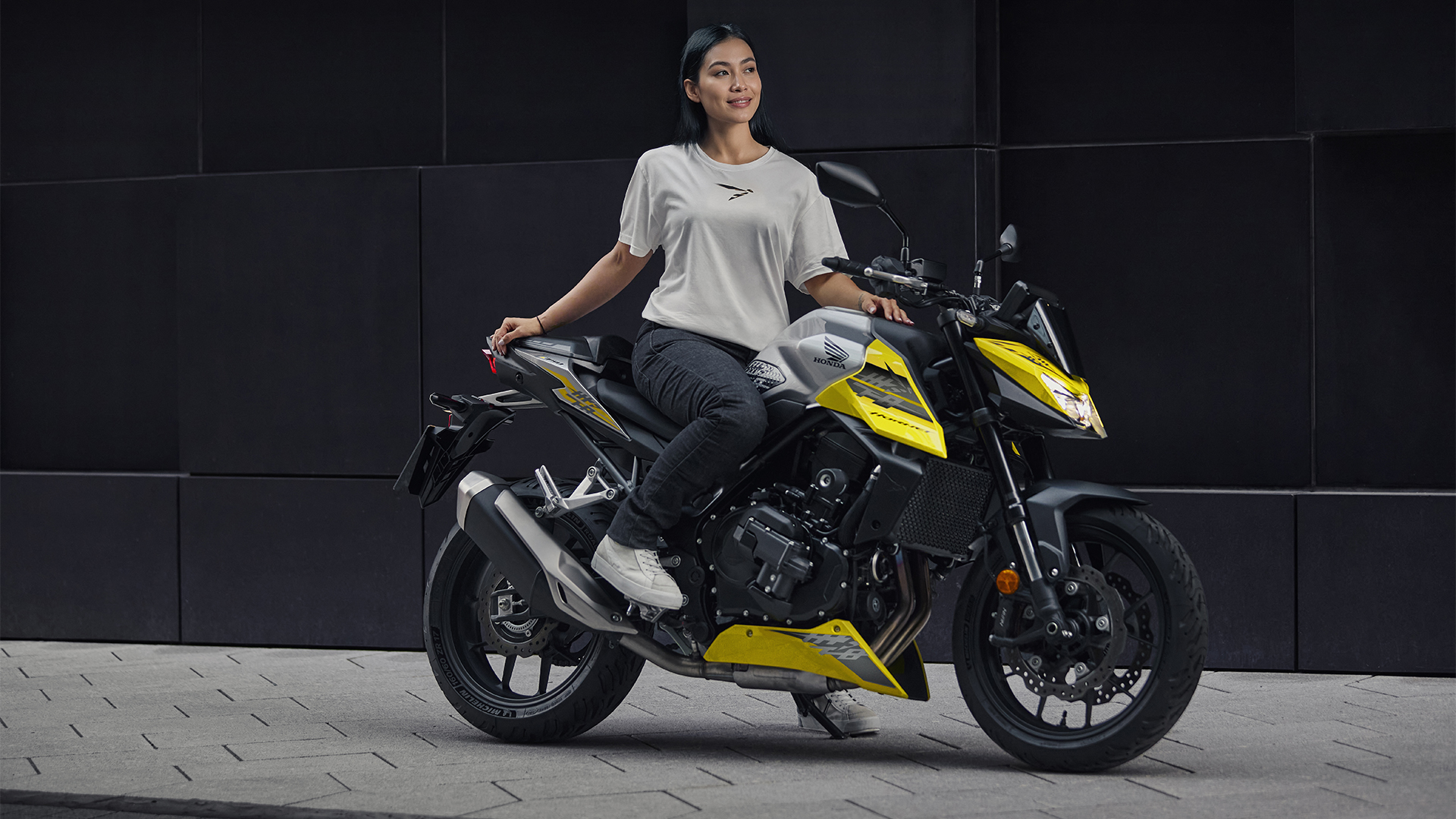 A yellow and black Honda CB300F motorcycle parked on a paved surface against a dark wall. A person is seated on the bike, wearing a white t-shirt and jeans.